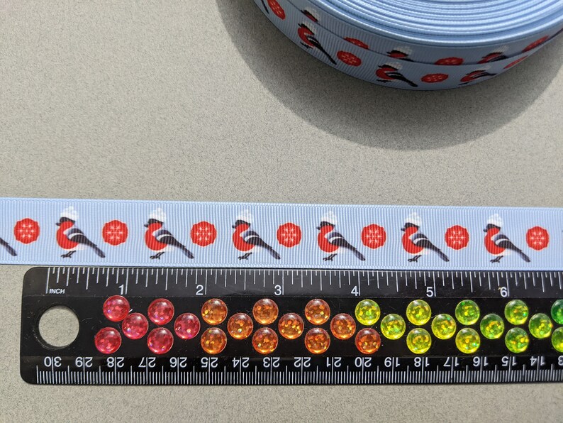 May include: A blue ribbon with a repeating pattern of red robins wearing white hats and red snowflakes. The ribbon is shown next to a ruler with inch markings.