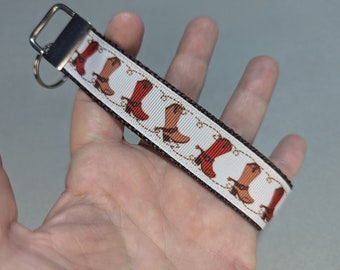 May include: A white and brown key chain with a silver key ring. The key chain has a repeating pattern of red cowboy boots.