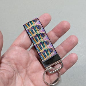 May include: A colorful keychain wrist strap with a repeating pattern of abstract shapes in shades of yellow, teal, purple, and black. The strap is attached to a silver-colored metal clasp and key ring.