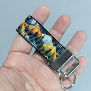 May include: A black and yellow patterned key chain with a silver metal key ring. The pattern features a mountain range with trees and a sunset.