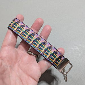 May include: A colorful wristlet key fob with a repeating pattern of stylized cupcakes in shades of purple, yellow, and teal. The key fob has a black background and a silver-colored metal clasp and key ring.