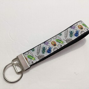 May include: A white and black key chain with a silver key ring. The white part of the key chain has a pattern of colorful bugs and insects.