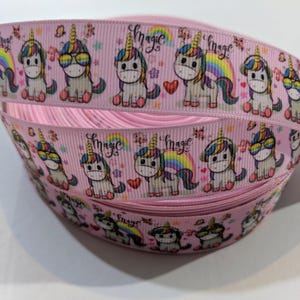 May include: A spool of light pink grosgrain ribbon featuring a repeating pattern of cartoon unicorns with colourful manes, rainbows, and hearts. The word magic is printed in a cursive script between the unicorn illustrations along the ribbon length.