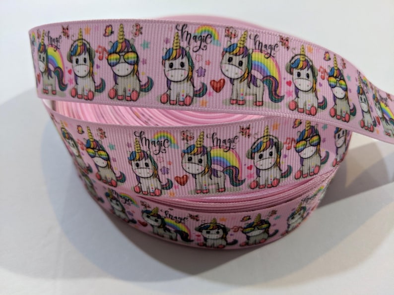 May include: A spool of light pink grosgrain ribbon featuring a repeating pattern of cartoon unicorns with colourful manes, rainbows, and hearts. The word magic is printed in a cursive script between the unicorn illustrations along the ribbon length.