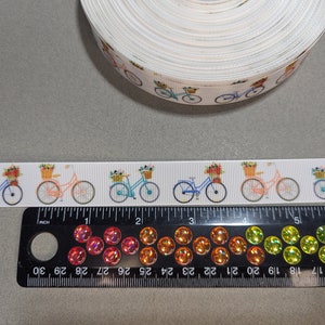 May include: White ribbon with a repeating pattern of bicycles with baskets of flowers. The ribbon is 1 inch wide and is shown next to a ruler.