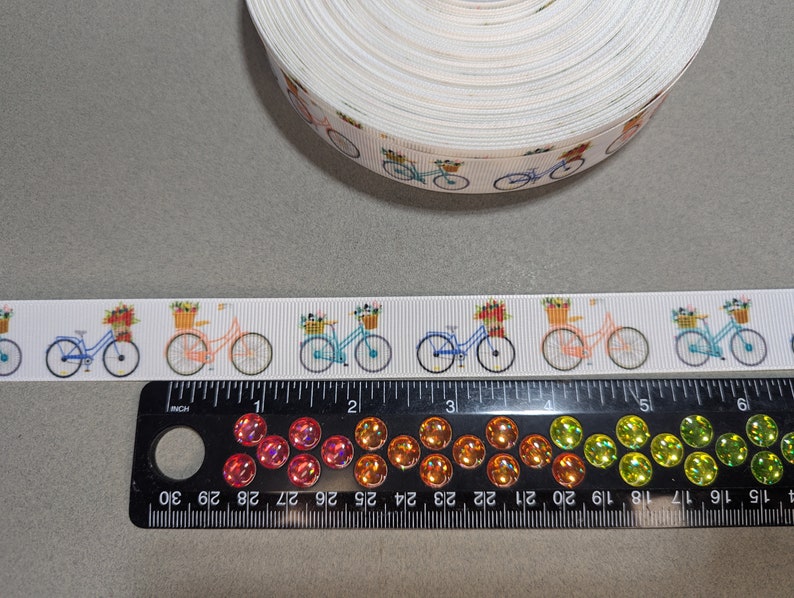 May include: White ribbon with a repeating pattern of bicycles with baskets of flowers. The ribbon is 1 inch wide and is shown next to a ruler.