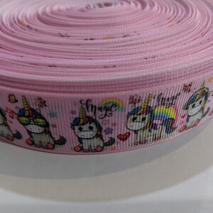May include: A roll of light pink grosgrain ribbon featuring a repeating pattern of cartoon unicorns, rainbows, hearts, and stars. The word magic is printed in a cursive black font above the unicorns, which are depicted with colourful manes.