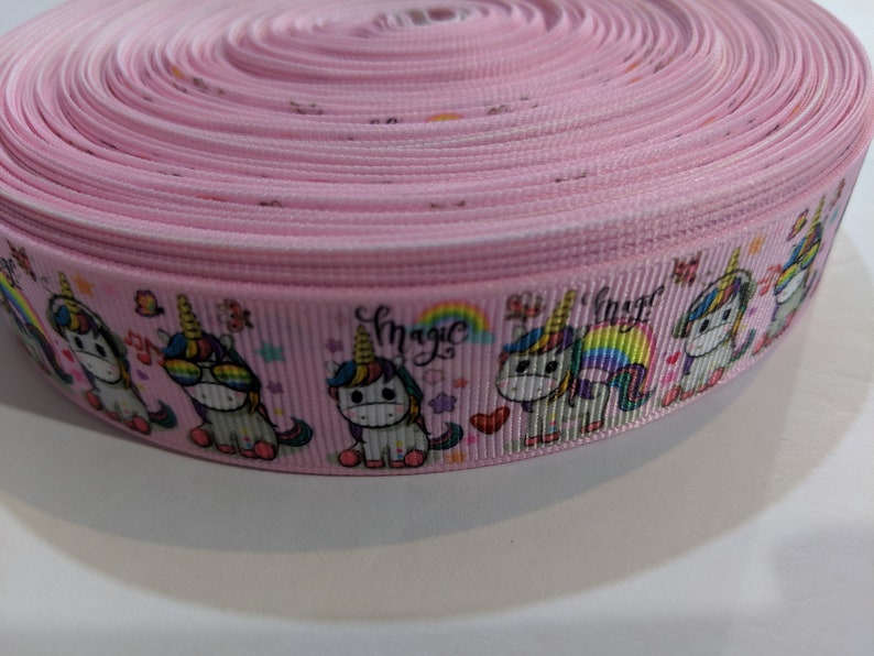 May include: A roll of light pink grosgrain ribbon featuring a repeating pattern of cartoon unicorns, rainbows, hearts, and stars. The word magic is printed in a cursive black font above the unicorns, which are depicted with colourful manes.