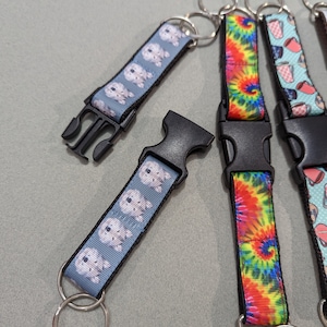 May include: Keychains with various patterned straps and black plastic buckles. Designs include a light blue strap with white dog illustrations, a tie-dye strap, and a strap with coffee cup illustrations. Each keychain has a silver metal ring.