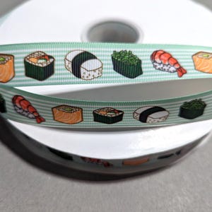 May include: A spool of light green grosgrain ribbon featuring a repeating pattern of various sushi pieces, including salmon nigiri, shrimp nigiri, and seaweed-wrapped rolls, printed in vibrant colors against a background of thin white horizontal stripes.