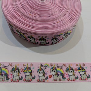 May include: A spool of light pink grosgrain ribbon featuring a repeating pattern of cartoon unicorns with colourful manes, rainbows, and the word magic in cursive script. The unicorns are depicted with various accessories including sunglasses.