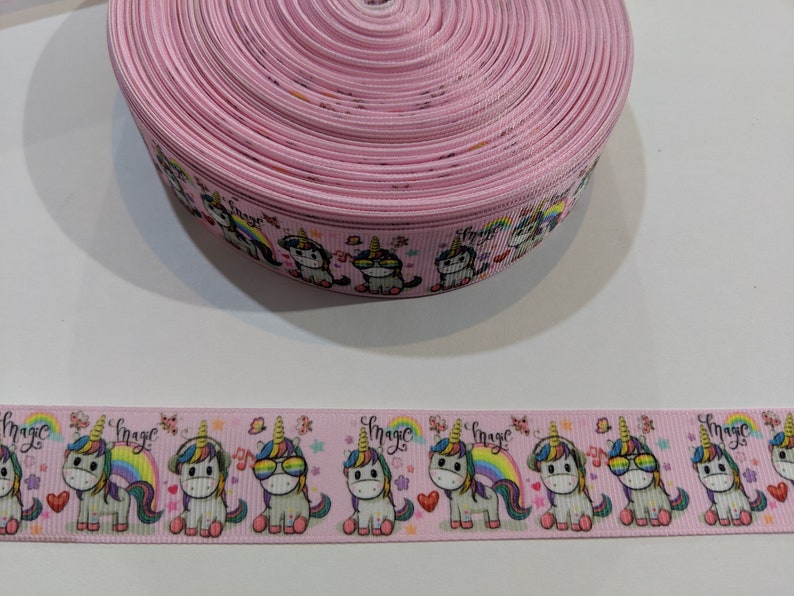 May include: A spool of light pink grosgrain ribbon featuring a repeating pattern of cartoon unicorns with colourful manes, rainbows, and the word magic in cursive script. The unicorns are depicted with various accessories including sunglasses.
