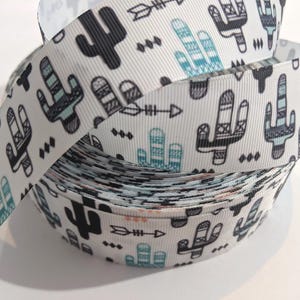 May include: A roll of white ribbon featuring a repeating pattern of black and teal cacti and arrows. The ribbon is tightly wound, showcasing the detailed design. The ribbon is suitable for crafting and gift wrapping.