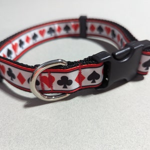 May include: A black and white dog collar with a red, white, and black pattern of playing card suits. The collar has a black plastic buckle and a silver metal D-ring.