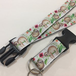 May include: A white fabric lanyard and a matching keychain featuring a repeating pattern of brown sloths, green tropical leaves, and small red flowers. Both items include a black plastic side-release buckle and a silver metal split ring.