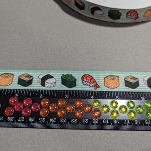 May include: A light green ribbon featuring a repeating pattern of various sushi rolls and shrimp nigiri. The ribbon is positioned above a black ruler that measures approximately 7 inches in length and features a row of colorful, iridescent cabochons.
