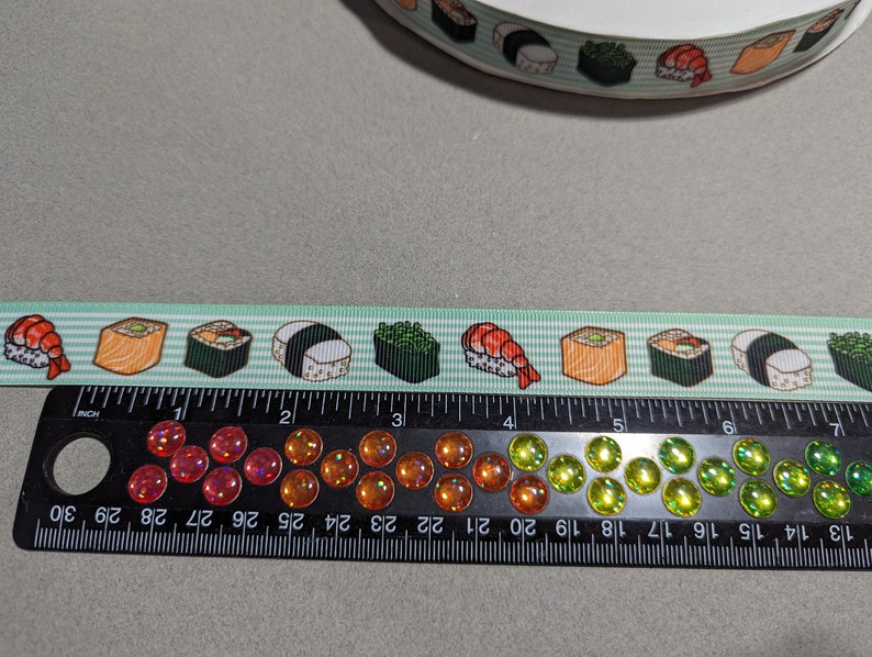 May include: A light green ribbon featuring a repeating pattern of various sushi rolls and shrimp nigiri. The ribbon is positioned above a black ruler that measures approximately 7 inches in length and features a row of colorful, iridescent cabochons.