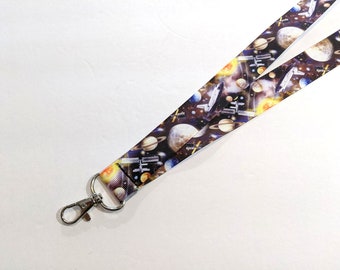 May include: A black and white lanyard with a space-themed print featuring planets, stars, and a spaceship. The lanyard has a silver metal clip.
