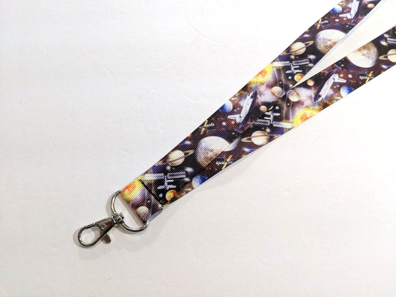 May include: A black and white lanyard with a space-themed print featuring planets, stars, and a spaceship. The lanyard has a silver metal clip.