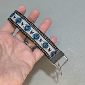 May include: A fabric keychain strap featuring a woven pattern of blue, black, and tan geometric shapes and small bull skull icons on a cream background, finished with a silver-toned metal clamp and a circular split ring for attaching keys.
