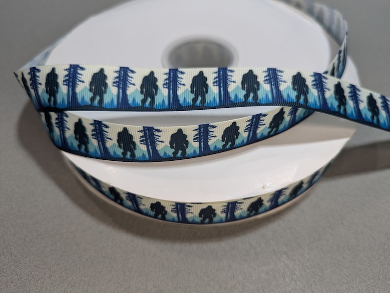 May include: A roll of white satin ribbon with a repeating pattern of black Bigfoot silhouettes walking through a blue and white forest.
