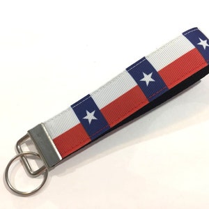 May include: A Texas flag-themed wristlet keychain. The fabric features the Texas flag design: a blue vertical stripe with a white star, white horizontal stripe, and red horizontal stripe. The wristlet has a black backing and a silver-colored metal clasp and key ring.