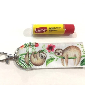 May include: A white and green sloth-themed key chain with a silver clip and a Carmex Classic Lip Balm tube. The lip balm tube is yellow with red lettering and says "Carmex Classic Lip Balm Medicated SPF 15".