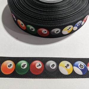 May include: Black ribbon featuring a repeating pattern of colorful billiard balls numbered 1 through 11. The ribbon is rolled up, with a section unrolled, displaying the full design. Ideal for crafting and embellishments.