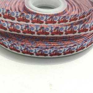 May include: A spool of white grosgrain ribbon featuring a repeating pattern of small red pickup trucks carrying blue snowflake designs. The ribbon is wound tightly around a central plastic core, displaying multiple layers of the printed fabric.