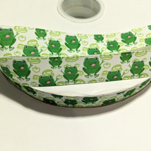 May include: White ribbon with a repeating pattern of green frogs and lily pads. The ribbon is rolled up on a white spool.