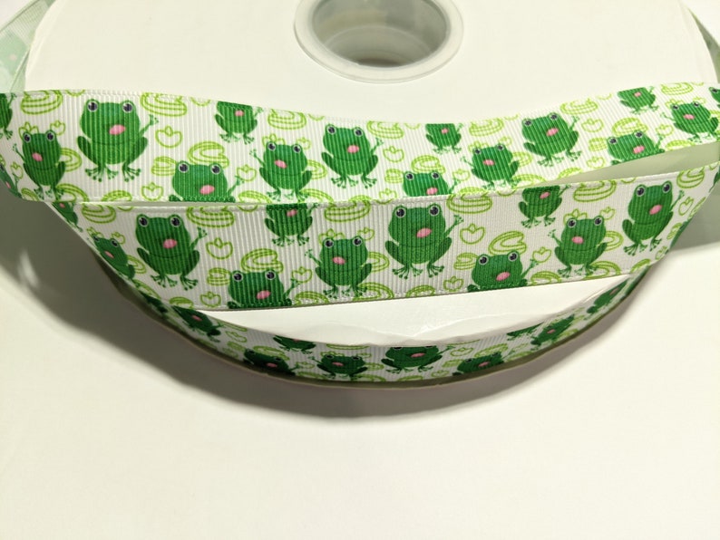 May include: White ribbon with a repeating pattern of green frogs and lily pads. The ribbon is rolled up on a white spool.