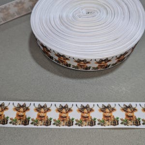 May include: White ribbon with a repeating pattern of brown and white Highland cows wearing bows. The ribbon is rolled up on a spool.