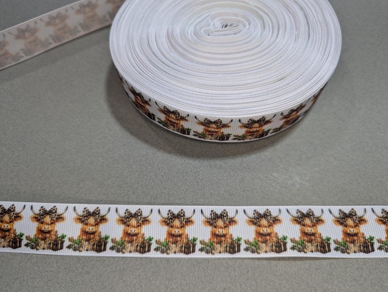 May include: White ribbon with a repeating pattern of brown and white Highland cows wearing bows. The ribbon is rolled up on a spool.