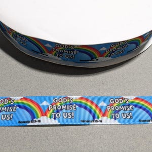 May include: A spool of light blue grosgrain ribbon featuring a repeating pattern of colorful rainbows, white clouds, and the black text "GOD'S PROMISE TO US!" followed by the scripture reference "Genesis 9:13-16" printed along the length of the fabric.