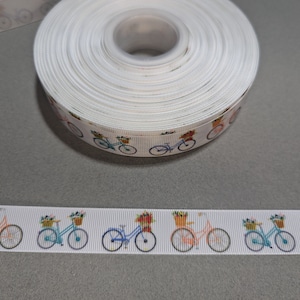 May include: White ribbon with a repeating pattern of colorful bicycles with baskets of flowers. The ribbon is rolled up and the pattern is visible on the side of the roll.