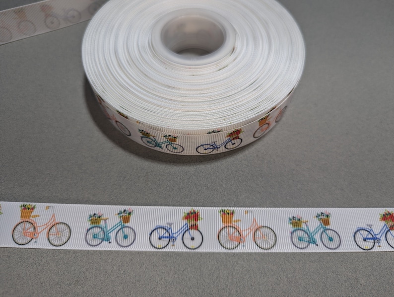 May include: White ribbon with a repeating pattern of colorful bicycles with baskets of flowers. The ribbon is rolled up and the pattern is visible on the side of the roll.