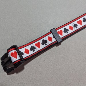 May include: A black dog collar with a white background and red and black playing card symbols. The collar has a black buckle.