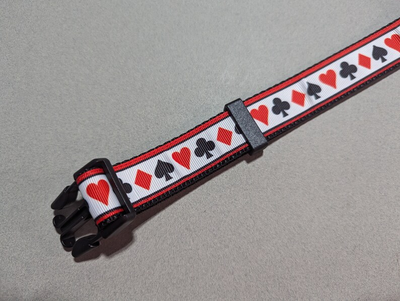 May include: A black dog collar with a white background and red and black playing card symbols. The collar has a black buckle.