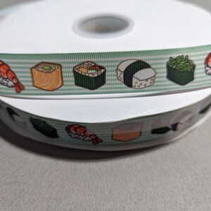 May include: A spool of light green and white striped ribbon featuring a repeating pattern of various sushi rolls and nigiri. The ribbon has a textured grosgrain finish and displays colorful illustrations of shrimp, seaweed, and rice-based sushi pieces.
