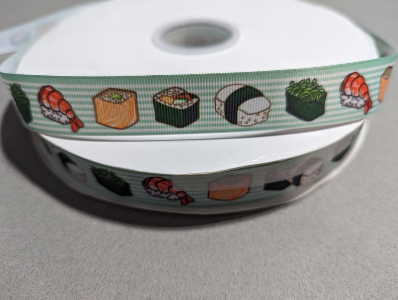 May include: A spool of light green and white striped ribbon featuring a repeating pattern of various sushi rolls and nigiri. The ribbon has a textured grosgrain finish and displays colorful illustrations of shrimp, seaweed, and rice-based sushi pieces.