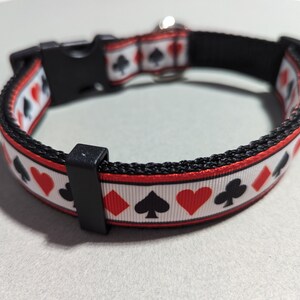 May include: A black and red dog collar with a white background and a repeating pattern of playing card suits. The collar has a black buckle and a silver D-ring.