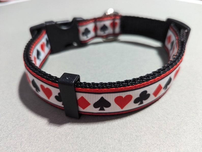 May include: A black and red dog collar with a white background and a repeating pattern of playing card suits. The collar has a black buckle and a silver D-ring.