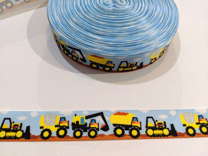 May include: Blue and white ribbon with a repeating pattern of yellow construction vehicles on a white background. The vehicles include a dump truck, a bulldozer, and a forklift.