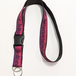 May include: A lanyard with a black strap and a pink and purple ribbon. The ribbon has the text "Be still and know that I am God" printed in black. The lanyard has a black plastic clasp and two silver key rings.