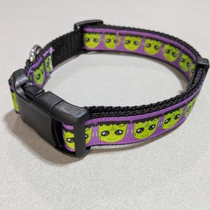May include: Black dog collar with a purple band featuring green monster faces. The collar has a black plastic buckle and a metal loop for attaching a leash. A fun and festive accessory for pets.