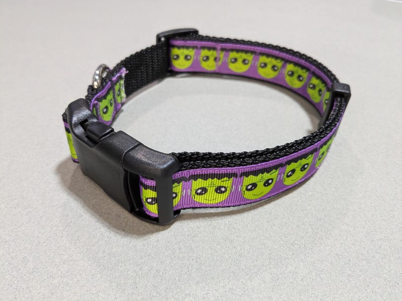 May include: Black dog collar with a purple band featuring green monster faces. The collar has a black plastic buckle and a metal loop for attaching a leash. A fun and festive accessory for pets.
