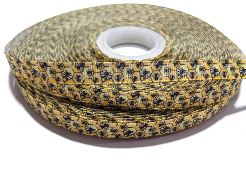 May include: A roll of decorative ribbon with a repeating pattern of yellow honeycombs and black bees. The ribbon is a light yellow colour with a white spool centre. The ribbon is likely used for crafts, gift wrapping, or other decorative purposes.