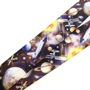 May include: A black and white space-themed lanyard with a repeating pattern of planets, stars, and a space shuttle. The lanyard is made of a woven fabric.
