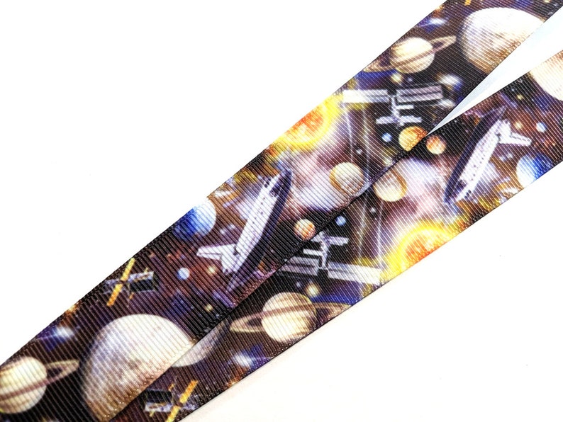 May include: A black and white space-themed lanyard with a repeating pattern of planets, stars, and a space shuttle. The lanyard is made of a woven fabric.