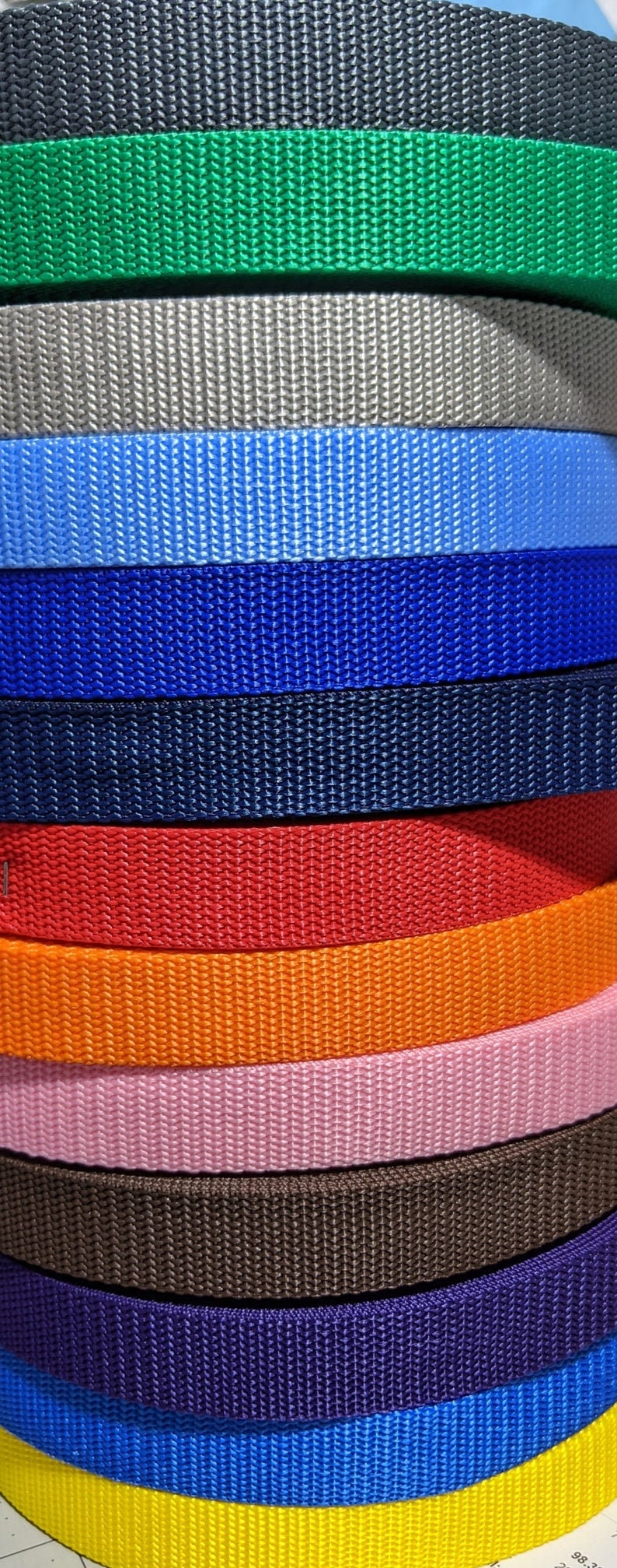 May include: A stack of colorful nylon webbing in various shades, including black, green, gray, blue, red, orange, pink, brown, and purple. The webbing has a tightly woven texture, suitable for straps or belts.